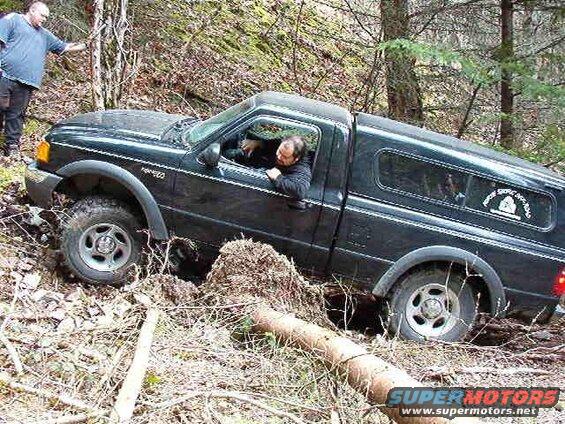 Black Ford pickup truck stuck in mud and trees, driver inside, another man outside.