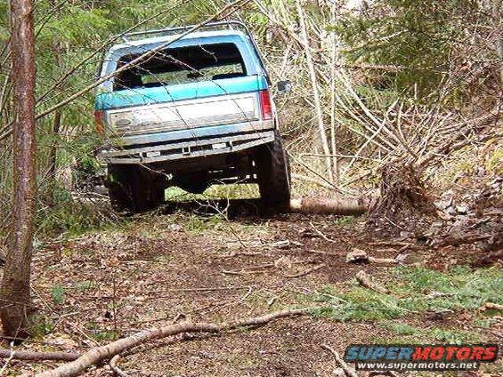 Blue Ford Bronco off-road, rear passenger side view, angled right, on uneven terrain with a log.