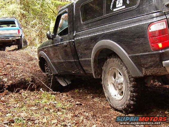 Black pickup truck driving off-road through a rutted dirt path with a blue pickup truck in the background.