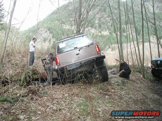 Brown Ford Bronco tilted on a steep, wooded trail with people nearby.