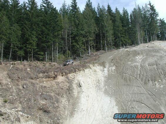 Silver SUV on the edge of a steep, eroded hillside with a forest behind it.