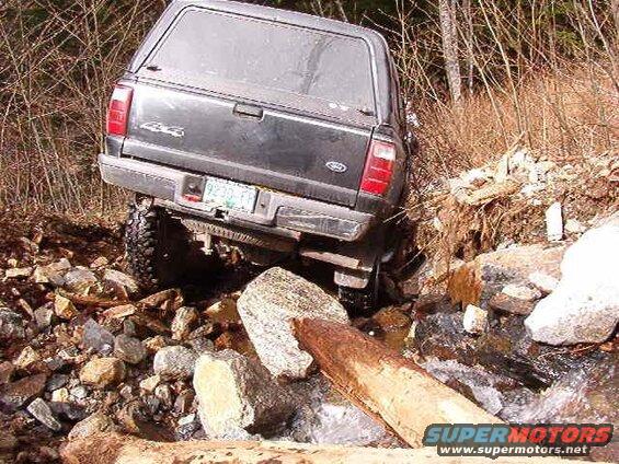 Dark-colored pickup truck stuck in a rocky stream with a log in the foreground.