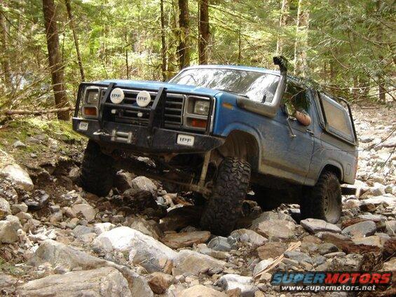 Blue 1985 Ford Bronco off-roading on a rocky trail in a forest.