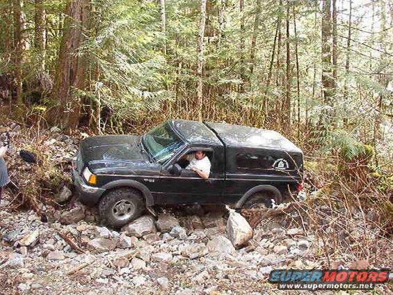 Black Ford pickup truck with camper shell on a rocky, wooded trail.