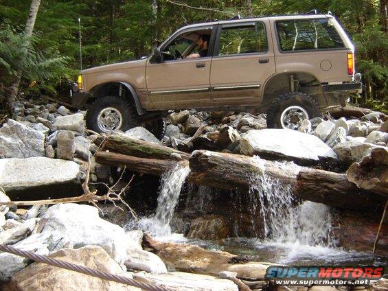 Tan Ford Explorer off-roading over logs and rocks in a stream.