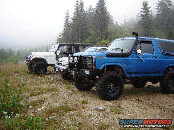 Blue 1985 Ford Bronco with snorkel, parked off-road with other vehicles.