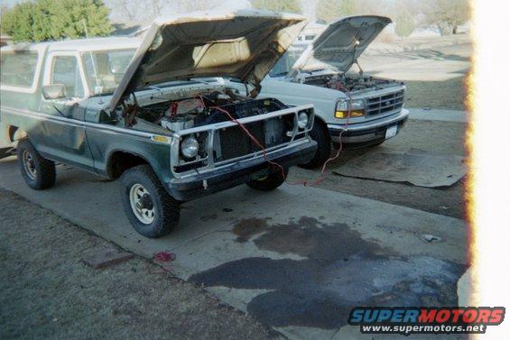 Green 1978 Ford Bronco with hood open, being jump-started by a white Ford F-Series truck