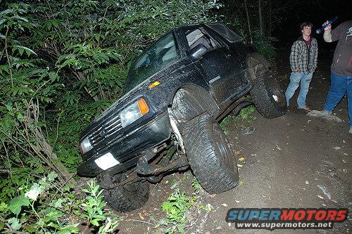 Black Suzuki Samurai tilted on wooded trail at night, two people nearby.