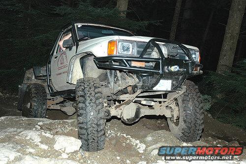 Modified white pickup truck on a rocky, wooded trail at night.