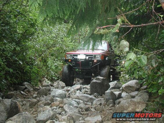 Red 4x4 off-road vehicle on a rocky trail surrounded by trees.