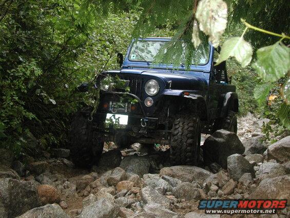 Blue Jeep CJ off-roading on a rocky trail with dense green trees.