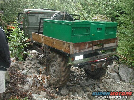 Green military truck with trailer and toolboxes on a rocky forest trail.