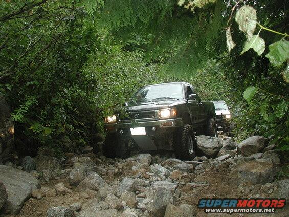 Two Toyota pickup trucks off-roading on a rocky, wooded trail.