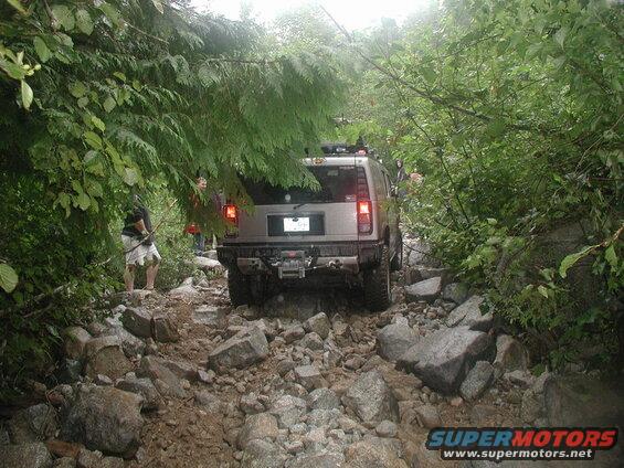 Silver Hummer H2 driving on a very rocky trail surrounded by dense green foliage.