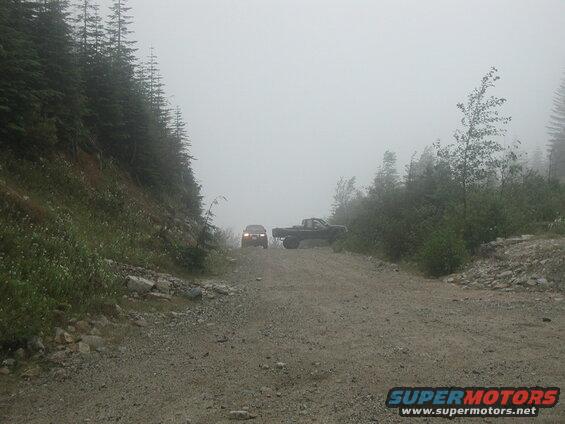 A dark SUV and a pickup truck on a foggy gravel road in a forest.