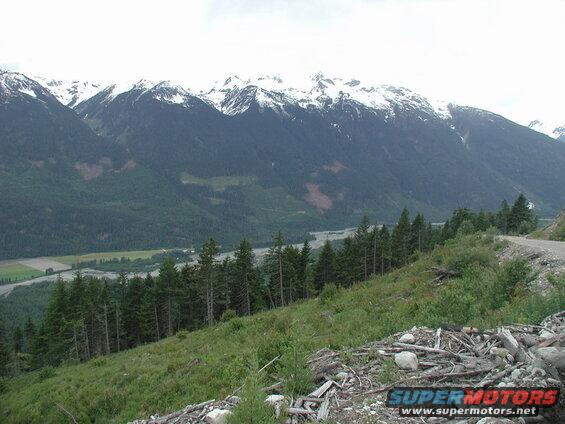 Mountain valley with river, snow-capped mountains, and a wooded slope with a dirt road.