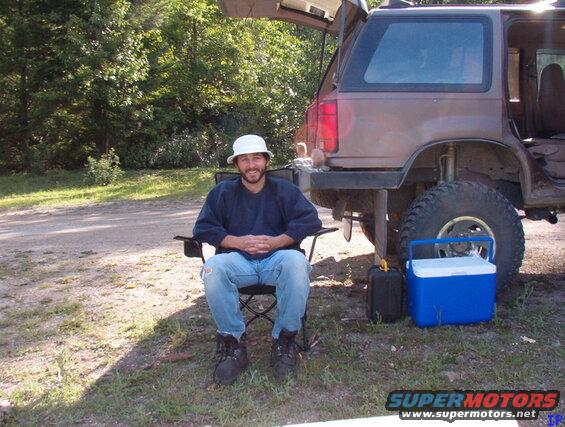 Man in camping chair next to lifted, muddy SUV with open hatch, cooler, and fuel can.