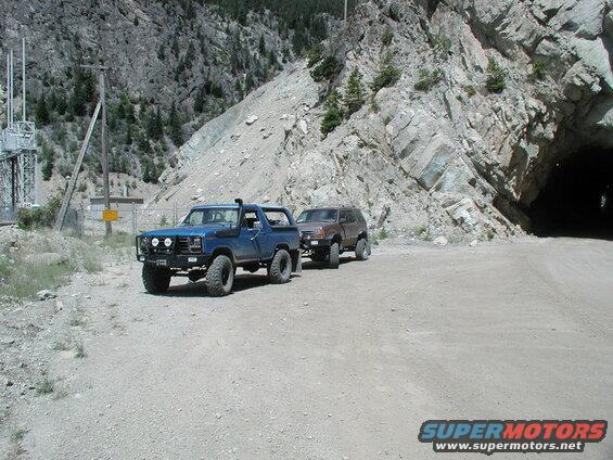 Lifted blue 1980s Ford Bronco parked on dirt road near tunnel entrance, with SUV and power substation.