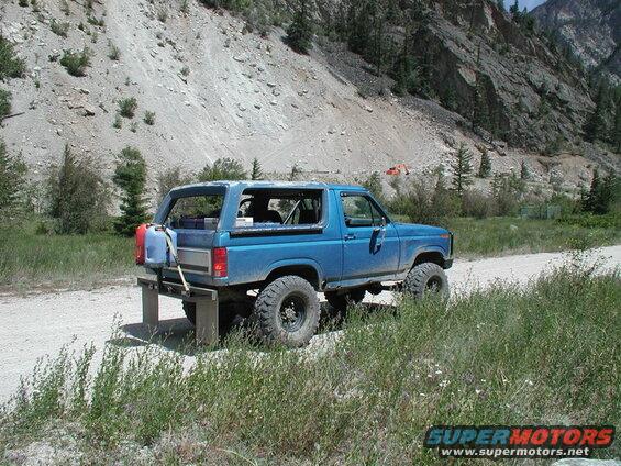 Lifted blue 1985 Ford Bronco parked on a dirt road with gas cans mounted on the back.