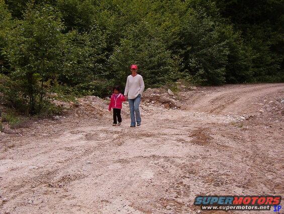 A woman and a young child walk on a dirt road in a wooded area.