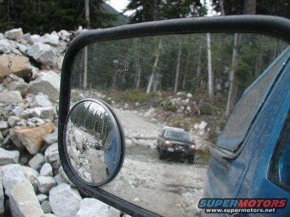 Reflection in a blue truck's side mirror shows another vehicle on a rocky, forested off-road path.