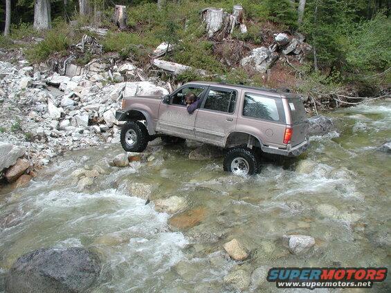 Lifted tan SUV driving through a rocky river.