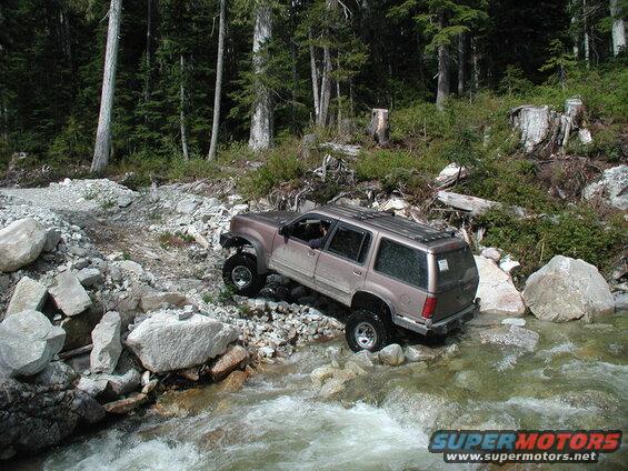 Tan SUV navigating a rocky stream in a forest.