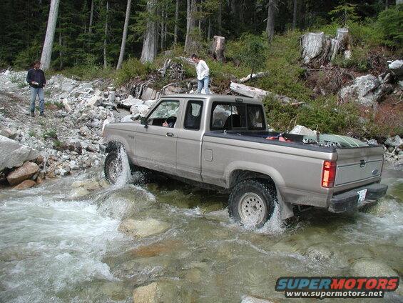 Tan Ford pickup truck driving through a rocky stream with two people nearby.