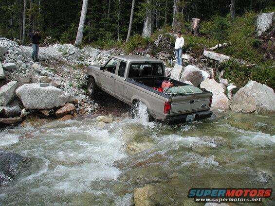 Tan Ford pickup truck driving through a rocky stream, with trees and boulders on the bank.