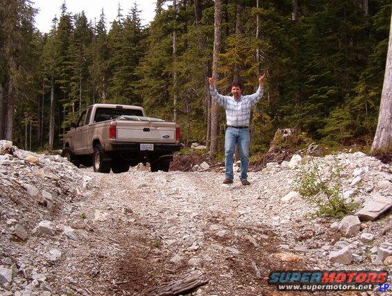Man on a rocky trail giving rock-on hand gesture next to a tan Ford pickup truck.