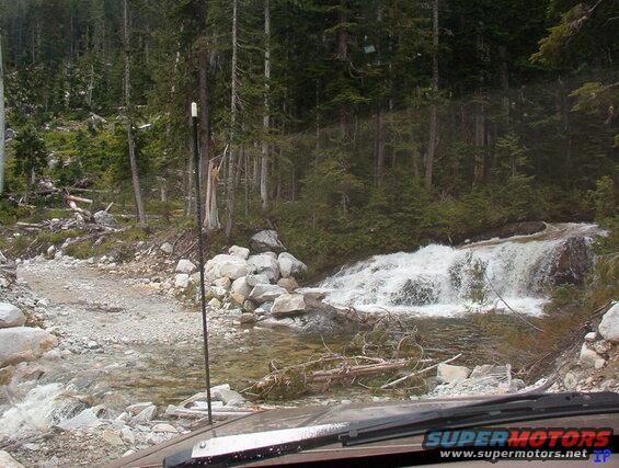 Front view of a vehicle on a gravel road near a waterfall in a forest.