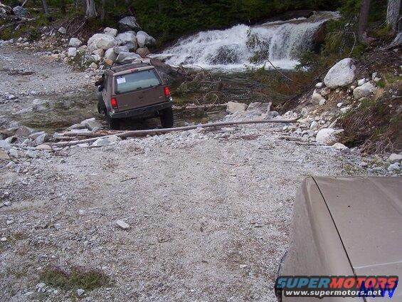 Brown SUV crossing a stream with a waterfall behind it. Another vehicle in foreground.