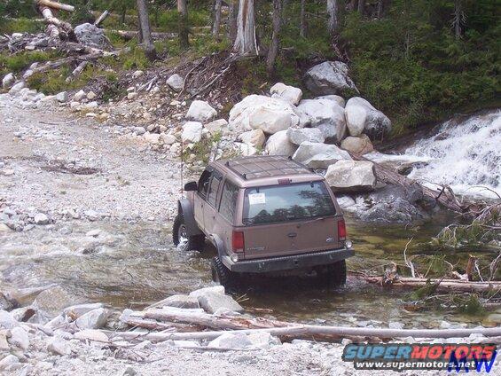 Brown SUV driving through a rocky stream near a waterfall.