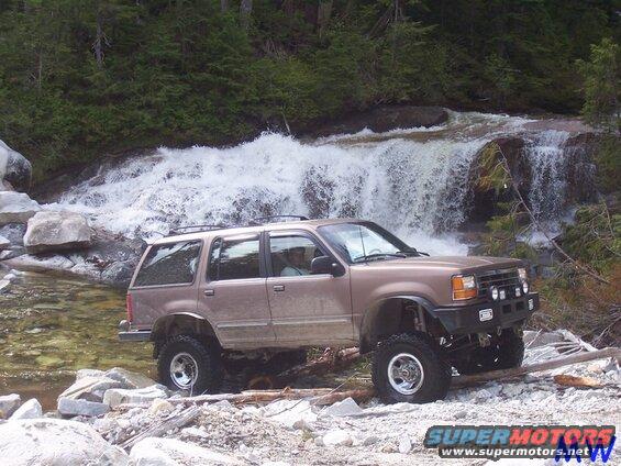 Lifted tan SUV driving through a stream near a waterfall.