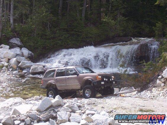 Tan Ford Explorer SUV with off-road modifications parked near a waterfall.