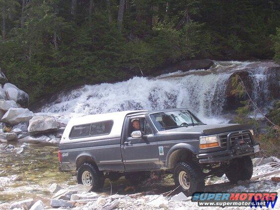 Dark gray pickup truck with white camper shell driving through a stream near a waterfall.