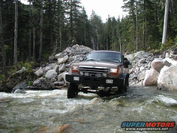 Lifted brown Ford Explorer driving through a river in a forest.