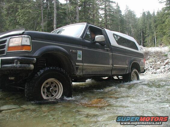 Dark Ford F-150 pickup truck with white camper shell driving through a stream in a forest.