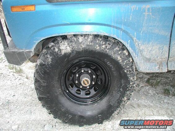 Blue 1985 Ford Bronco front wheel and fender with off-road tire covered in mud.