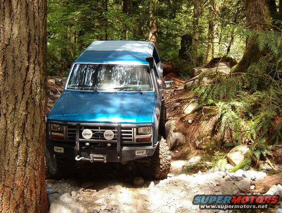 Blue 1980s Ford Bronco off-roading on a rocky forest trail.