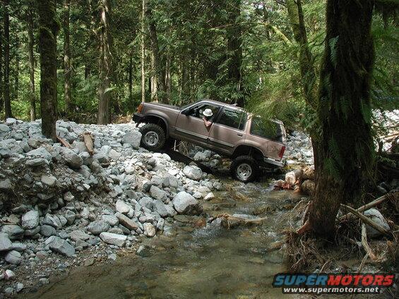Lifted tan Ford Explorer driving through a rocky stream in a forest.