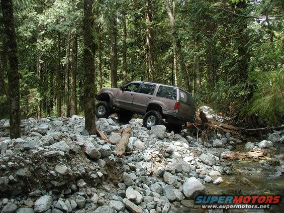 Lifted tan SUV driving over rocks in a forest next to a stream.