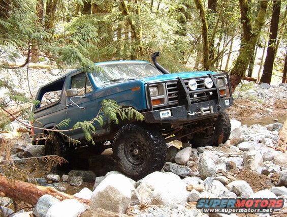 Blue Ford Bronco off-roading through a rocky stream in a forest.
