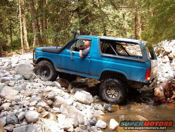 Blue 1980s Ford Bronco off-roading through a rocky stream with a snorkel.