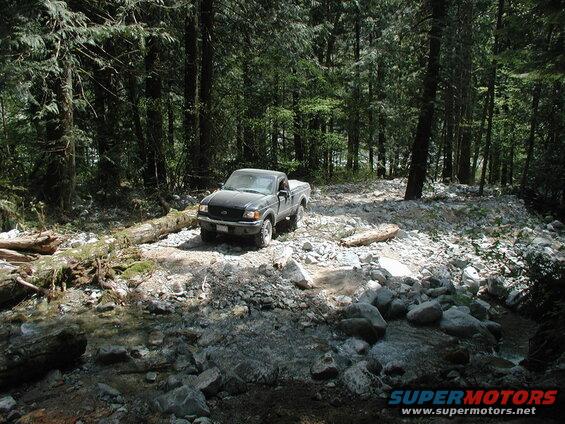 Dark pickup truck on a rocky forest trail next to a fallen log and stream.