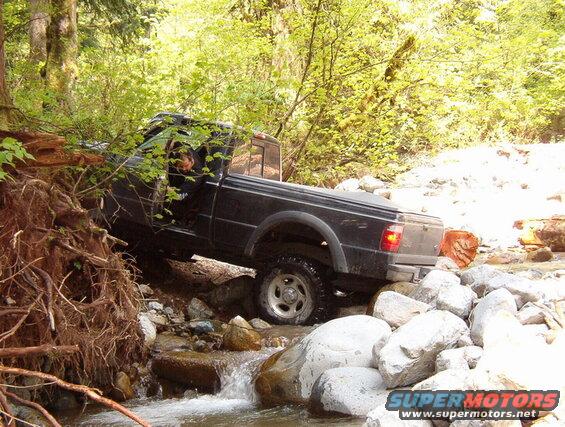 Black pickup truck driving through a rocky stream in a forest.