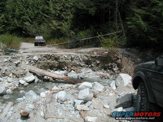 Dark SUV on a washed-out road with a stream and forest.