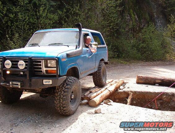 Blue Ford Bronco off-roading over logs across a trench.