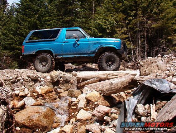 Blue 1980s Ford Bronco off-roading over logs and rocks in a forest.