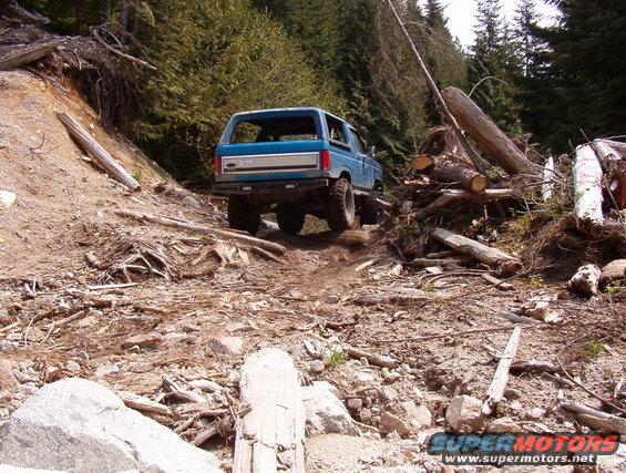 Blue 1985 Ford Bronco driving on a rough, wooded mountain trail.